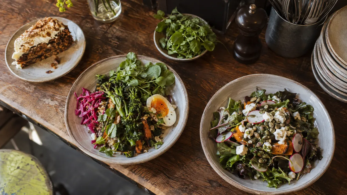 Plated salads and a slice of cake on a rustic cafe counter beside fresh herbs.