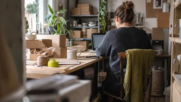 A shop owner checks a laptop beside stock boxes in a small homeware shop office.